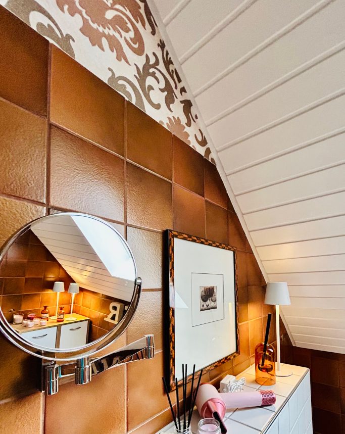 Bad 2 Wand mit braunen Fliesen, dekorativem Muster und Spiegel, dazu ein Bild und Lampe.