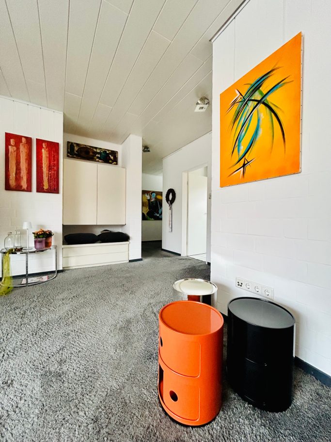 Wohnraum 4 Modernes Zimmer mit orangefarbenem Kunstwerk, farbigen Hockern und minimalistischer Einrichtung.