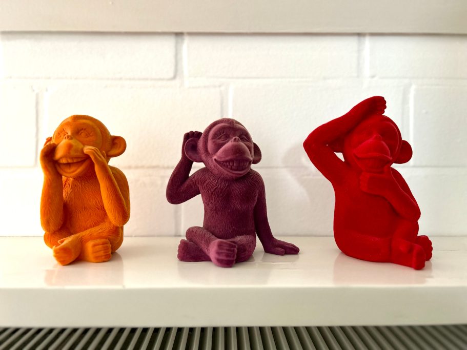Deko Drei bunte Affenskulpturen in Orange, Lila und Rot, sitzen auf einer weißen Fläche.