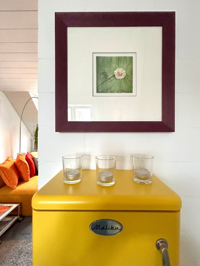 Malibu Schränkchen Eine gelbe Kommode mit einem gerahmten Bild und Gläsern darauf, im Hintergrund ein farbenfrohes Sofa.