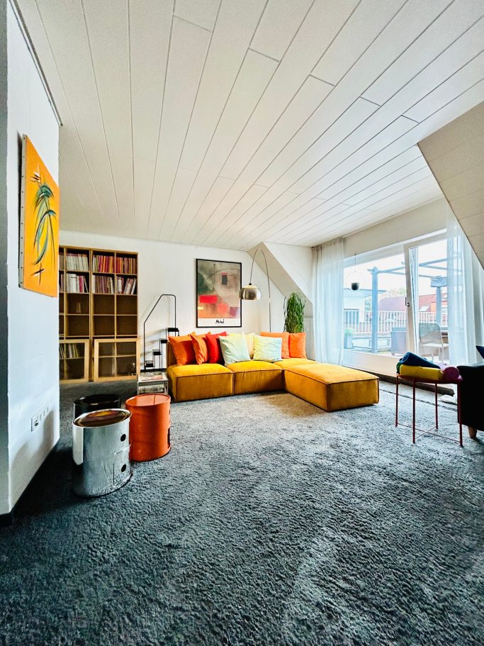 Wohnraum Helles Wohnzimmer mit gelbem Sofa, bunten Kissen und großen Fenstern.